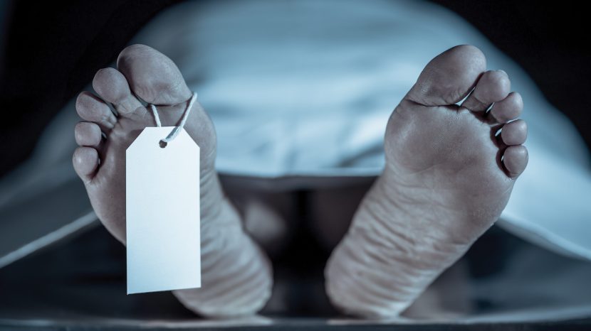 Close-up of dead body feet at morgue or hospital with toe label or information ring and identification blank tag. Cadaver lying on steal table covered with sheet on autopsy table. Death concept.